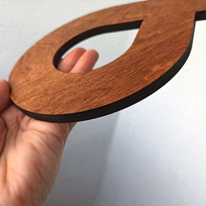May include: A wooden cutout of the letter "O" with a smaller cutout in the shape of a teardrop. The cutout is stained a dark brown colour and has a black border.