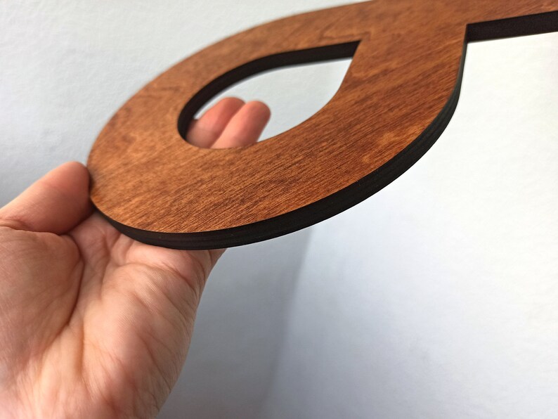 May include: A wooden cutout of the letter "O" with a smaller cutout in the shape of a teardrop. The cutout is stained a dark brown colour and has a black border.