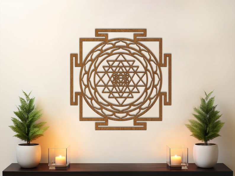 Op de afbeelding: Een bruine houten wanddecoratie met een geometrisch ontwerp, met een Sri Yantra, een heilig symbool in het hindoe&iuml;sme. De wanddecoratie is te zien op een witte muur met twee kleine potplanten en kaarsen op een houten plank.