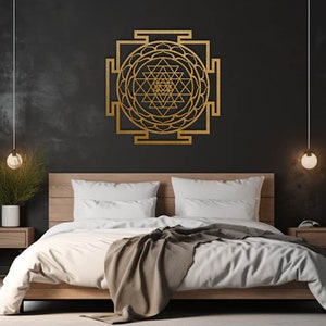 Op de afbeelding: Een gouden metalen wanddecoratie met een geometrisch ontwerp met een mandala-achtig patroon. De decoratie hangt aan een zwarte muur boven een houten bedframe met witte beddengoed.