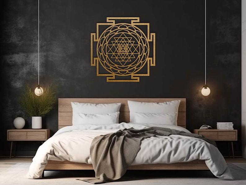 Op de afbeelding: Een gouden metalen wanddecoratie met een geometrisch ontwerp met een mandala-achtig patroon. De decoratie hangt aan een zwarte muur boven een houten bedframe met witte beddengoed.