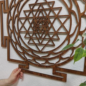 Op de afbeelding: Een bruine houten wanddecoratie met een geometrisch ontwerp. Het ontwerp is een Sri Yantra, een heilig symbool in het hindoe&iuml;sme. De wanddecoratie is gemaakt van hout en heeft een donkerbruine afwerking.