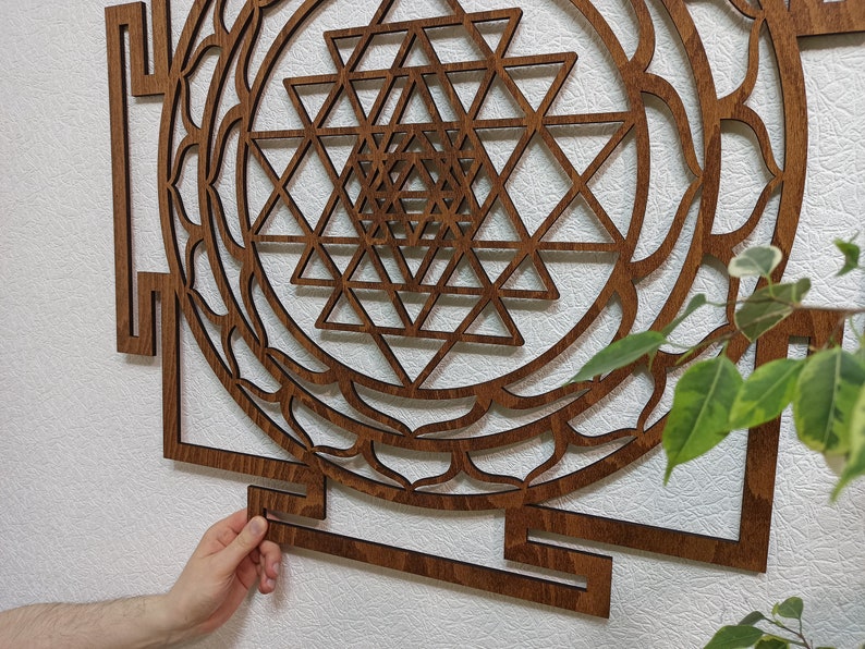 Op de afbeelding: Een bruine houten wanddecoratie met een geometrisch ontwerp. Het ontwerp is een Sri Yantra, een heilig symbool in het hindoe&iuml;sme. De wanddecoratie is gemaakt van hout en heeft een donkerbruine afwerking.