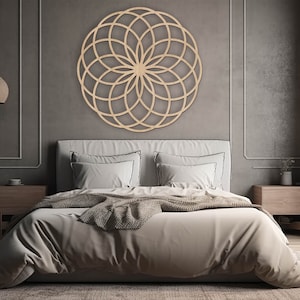 K&ouml;nnte beinhalten: Eine geometrische Mandala-Wanddekoration aus Holz mit einem runden Design in einem Schlafzimmer. Das Mandala h&auml;ngt &uuml;ber einem Bett mit wei&szlig;en Bettw&auml;sche und einer braunen Wolldecke.