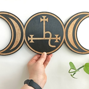 Op de afbeelding: Houten wanddecoratie met een drievoudige maansikkel symbool met een zwarte achtergrond en een gouden zegel in het midden. De zegel is een gestileerd kruis met een slang eromheen gewikkeld.