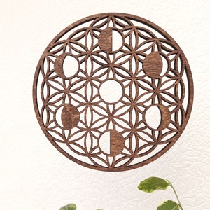 Op de afbeelding: Een ronde houten wanddecoratie met een lasergesneden ontwerp van de maanfasen in een bloem des levens patroon. Het hout heeft een warme bruine kleur en het ontwerp is ingewikkeld en gedetailleerd.