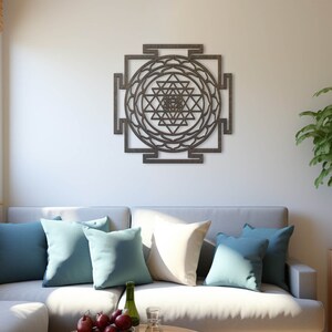 Op de afbeelding: Een grijze bank met blauwe en witte kussens in een woonkamer met een houten wandsculptuur van een Sri Yantra mandala, een kamerplant en een raam met jaloezie&euml;n.