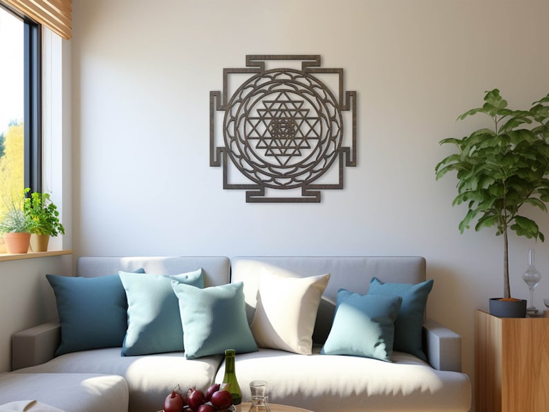 Op de afbeelding: Een grijze bank met blauwe en witte kussens in een woonkamer met een houten wandsculptuur van een Sri Yantra mandala, een kamerplant en een raam met jaloezie&euml;n.