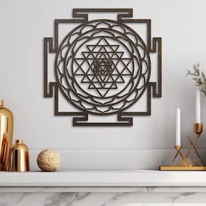 Op de afbeelding: Een bruine houten wanddecoratie met een geometrisch ontwerp. Het ontwerp is een Sri Yantra, een heilig symbool in het hindoe&iuml;sme. De wanddecoratie hangt aan een witte muur met een plank eronder.