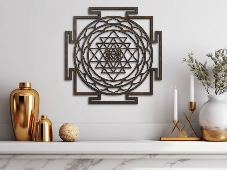 Op de afbeelding: Een bruine houten wanddecoratie met een geometrisch ontwerp. Het ontwerp is een Sri Yantra, een heilig symbool in het hindoe&iuml;sme. De wanddecoratie hangt aan een witte muur met een plank eronder.