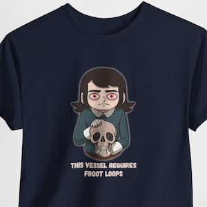 Op de afbeelding: Marineblauw t-shirt met een cartoon illustratie van een persoon die een schedel vasthoudt. De persoon heeft rode ogen en draagt een blauw shirt. De tekst op het shirt luidt "THIS VESSEL REQUIRES FROOT LOOPS".