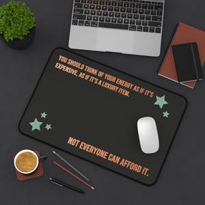 May include: Black rectangular mousepad with the text "YOU SHOULD THINK OF YOUR ENERGY AS IF IT'S EXPENSIVE, AS IF IT'S A LUXURY ITEM. NOT EVERYONE CAN AFFORD IT." and teal stars. A white mouse sits on the pad.