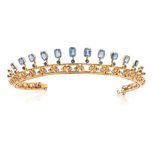May include: A gold tiara featuring a curved band with an intricate openwork design. The piece is adorned with a row of upright, cushion-cut light blue sapphires, each accented by a smaller round blue sapphire at the base and clear white diamonds.