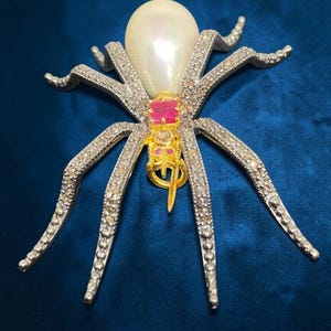 May include: A spider-shaped brooch featuring a large, iridescent white pearl as the abdomen. The thorax is accented with a square-cut red gemstone and smaller red stones. The eight legs are encrusted with numerous small, clear pavé-set stones.