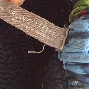 Może przedstawiać: Zbliżenie na szarą metkę z tekstem "URBAN OUTFITTERS" i "Made in India/Fabriqu&eacute; en Inde". Metka jest przymocowana do ciemnoniebieskiego ubrania z niebiesko-zielonym nadrukiem kwiatowym.