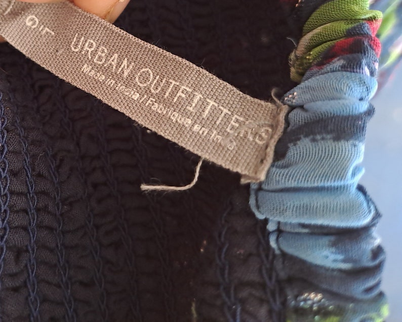 Może przedstawiać: Zbliżenie na szarą metkę z tekstem "URBAN OUTFITTERS" i "Made in India/Fabriqu&eacute; en Inde". Metka jest przymocowana do ciemnoniebieskiego ubrania z niebiesko-zielonym nadrukiem kwiatowym.
