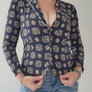 May include: A navy blue blouse with a floral pattern in beige and white. The long-sleeved blouse has a ruffled trim along the neckline. The model is wearing blue jeans.