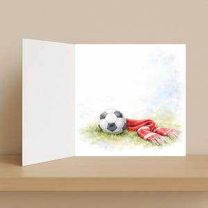以下が含まれることがあります： サッカーボールと、白いストライプの赤いスカーフの水彩イラストが描かれたグリーティングカード。ボールは白と黒で、緑の草の上に置かれています。カードは開いており、内側は空白です。