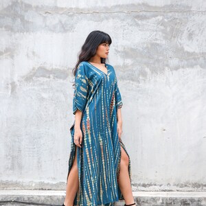 May include: A woman wearing a long, teal blue maxi dress with a tie-dye pattern. The dress has side slits and a V-neckline. She is wearing black heeled sandals.