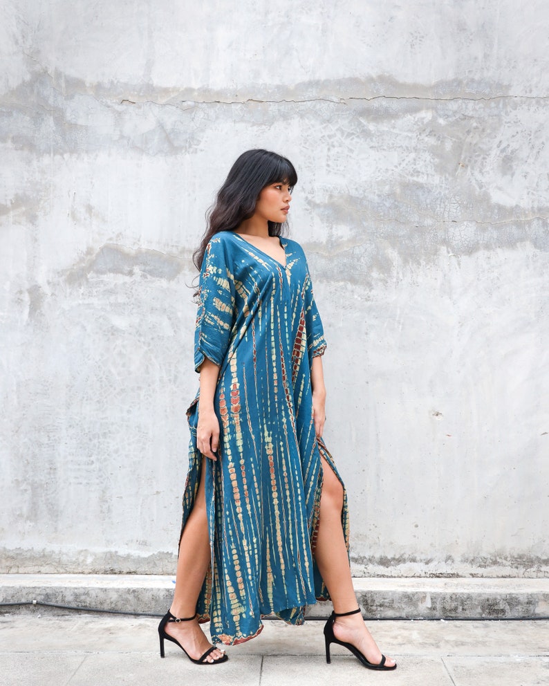 May include: A woman wearing a long, teal blue maxi dress with a tie-dye pattern. The dress has side slits and a V-neckline. She is wearing black heeled sandals.