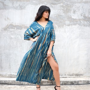 May include: A woman wearing a long, teal blue tie-dye dress with a V-neckline and short sleeves. She is standing in front of a concrete wall and is wearing black heels.