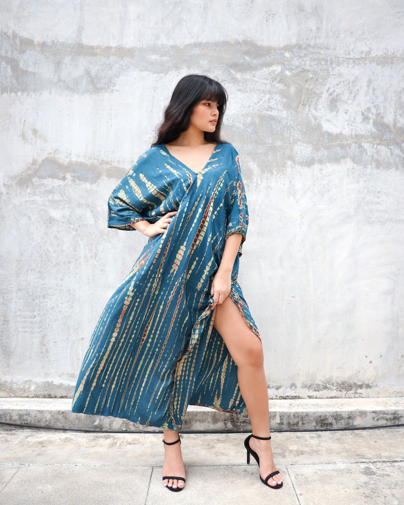 May include: A woman wearing a long, teal blue tie-dye dress with a V-neckline and short sleeves. She is standing in front of a concrete wall and is wearing black heels.