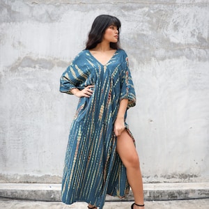 May include: A long, teal blue maxi dress with a tie-dye pattern in shades of brown, green, and white. The dress has a V-neckline and three-quarter sleeves. The model is wearing black heeled sandals.