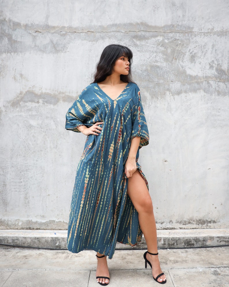 May include: A long, teal blue maxi dress with a tie-dye pattern in shades of brown, green, and white. The dress has a V-neckline and three-quarter sleeves. The model is wearing black heeled sandals.