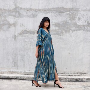 May include: A woman wearing a long, blue and white tie-dye dress with a V-neck and a side slit. She is wearing black heeled sandals and standing in front of a concrete wall.