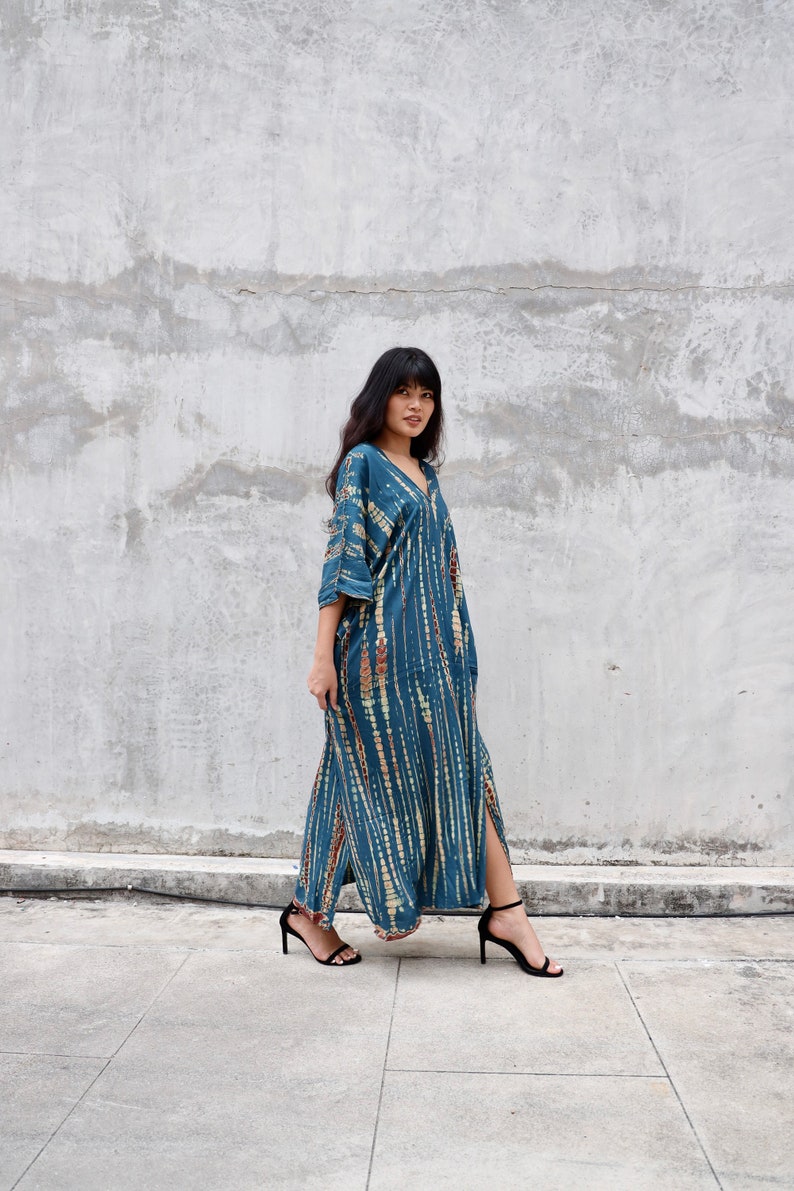 May include: A woman wearing a long, blue and white tie-dye dress with a V-neck and a side slit. She is wearing black heeled sandals and standing in front of a concrete wall.