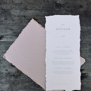 May include: A white dinner menu with torn edges, featuring the text "M&G DINNER" and a list of courses and ingredients. A pale pink envelope is beneath the menu, all on a dark wood surface.
