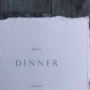 May include: A close-up of a white paper with a torn edge. The word "DINNER" is printed in a dark green font, with the initials "M&G" above it. A horizontal line is below the word "DINNER".