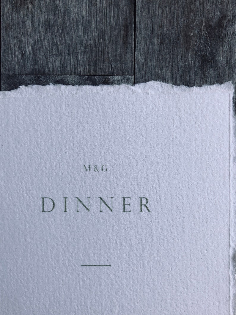 May include: A close-up of a white paper with a torn edge. The word "DINNER" is printed in a dark green font, with the initials "M&G" above it. A horizontal line is below the word "DINNER".