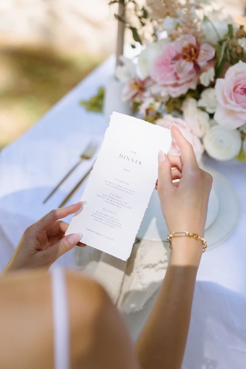 May include: A white menu card with the word "DINNER" at the top, held by a person. The menu has a torn edge and features a list of courses. A gold bracelet is visible on the person's wrist. A floral arrangement is in the background.