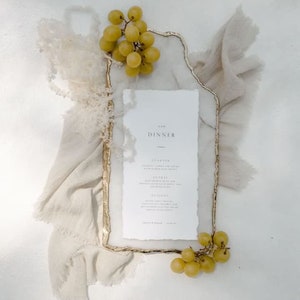 May include: A dinner menu with the word "DINNER" at the top, framed by a gold-edged rectangle. The menu is surrounded by a beige cloth and green grapes. The menu includes the words "STARTERS", "ENTREES", and "DESSERT".