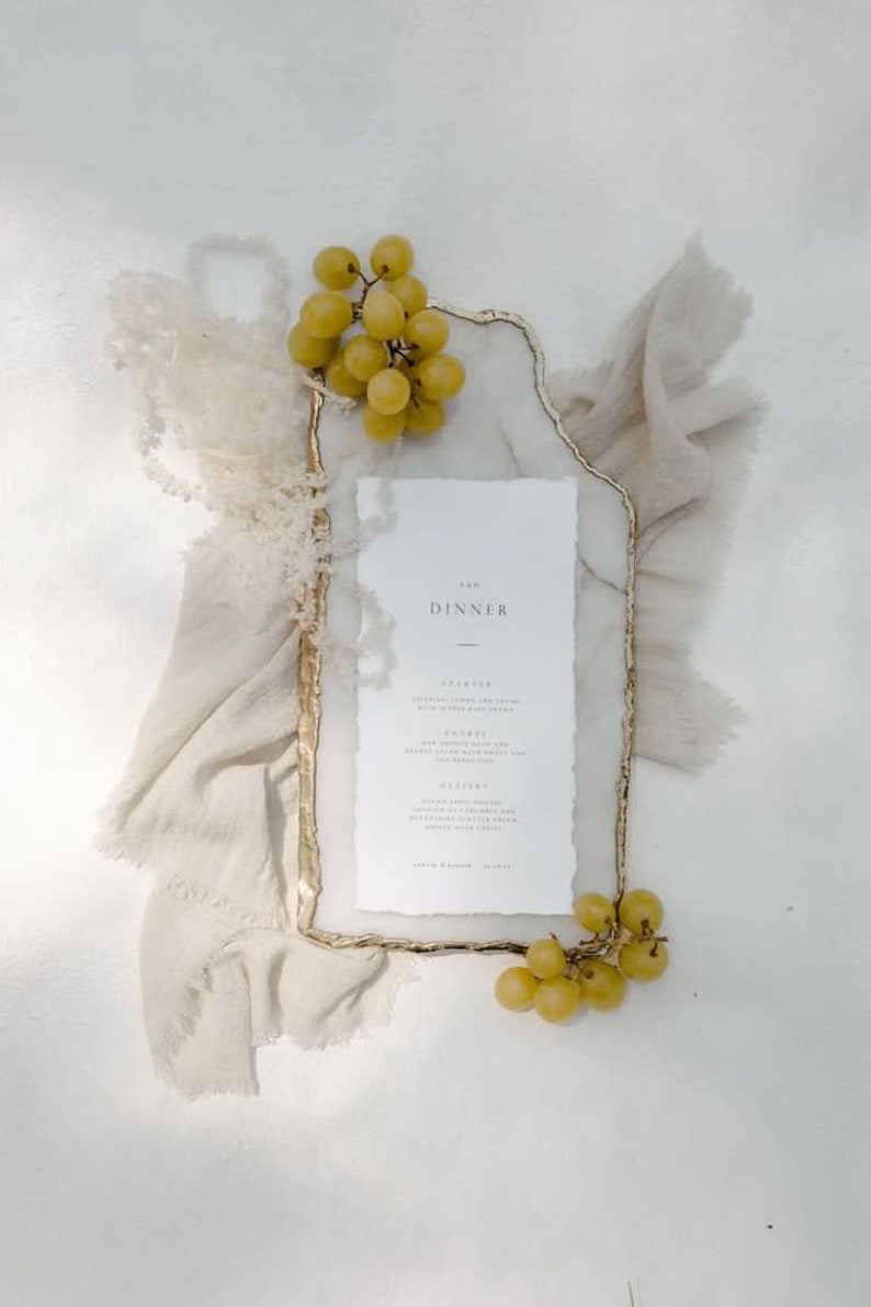 May include: A dinner menu with the word "DINNER" at the top, framed by a gold-edged rectangle. The menu is surrounded by a beige cloth and green grapes. The menu includes the words "STARTERS", "ENTREES", and "DESSERT".