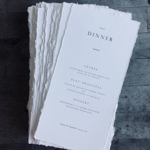 May include: A stack of white paper dinner menus with deckled edges. The menus feature the text "L&L DINNER" at the top, followed by menu items in French. The bottom of the menu reads "laura & layachi 03.07.21".
