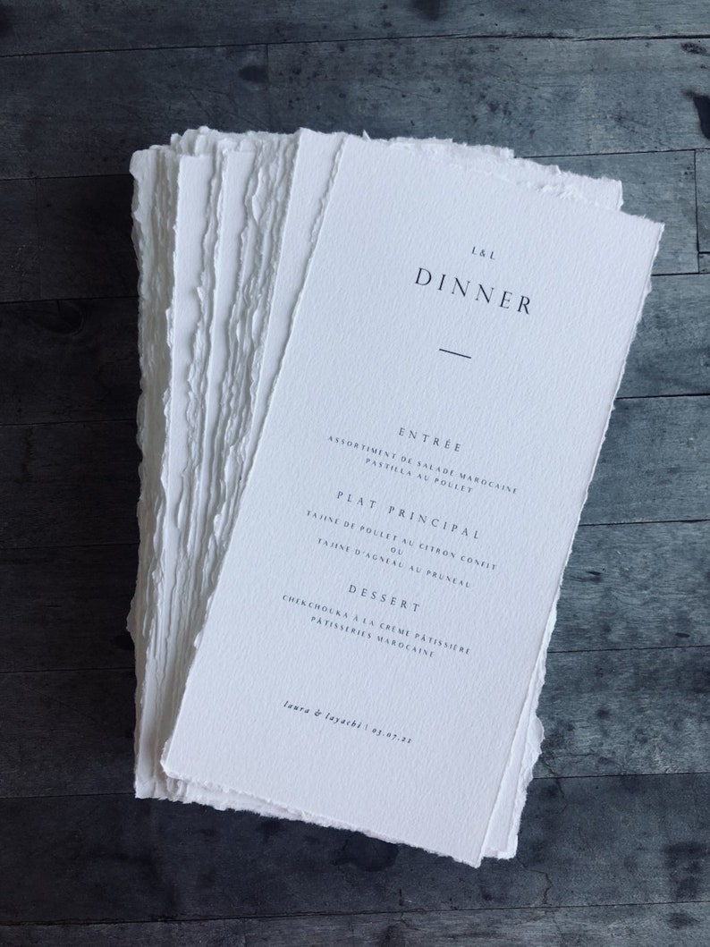 May include: A stack of white paper dinner menus with deckled edges. The menus feature the text "L&L DINNER" at the top, followed by menu items in French. The bottom of the menu reads "laura & layachi 03.07.21".