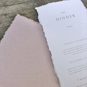 May include: A close-up of a dinner menu and a pink paper. The menu is white with the word "DINNER" in a dark gray font. The menu has a torn edge. The pink paper has a torn edge.