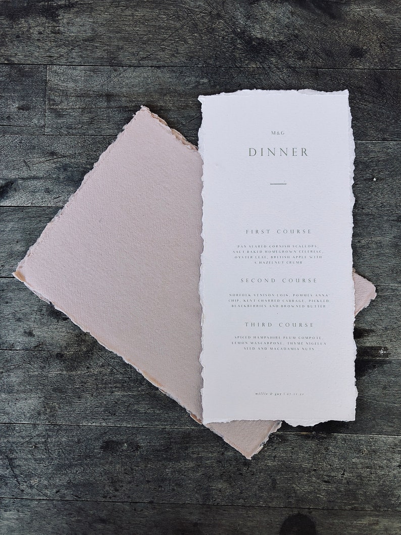 May include: A white dinner menu with torn edges, featuring the text "M&G DINNER" and a list of courses and ingredients. A pale pink envelope is beneath the menu, all on a dark wood surface.
