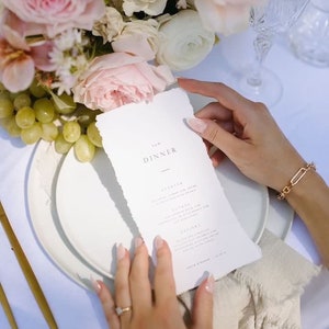 May include: A formal dinner setting with a menu, flowers, and grapes. The menu reads "The Dinner" with a list of courses. A gold bracelet and ring are visible. Pink and white roses and other flowers are in the background.