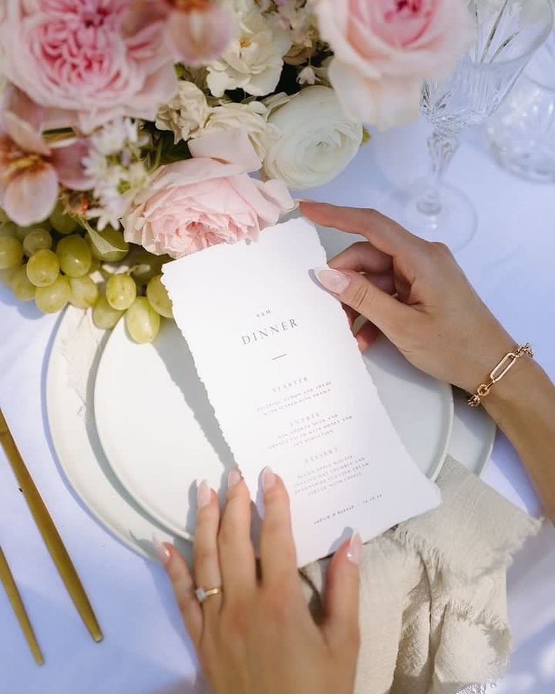 May include: A formal dinner setting with a menu, flowers, and grapes. The menu reads "The Dinner" with a list of courses. A gold bracelet and ring are visible. Pink and white roses and other flowers are in the background.