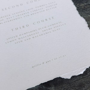 May include: A close-up of a white paper menu with a torn edge. The menu lists three courses with detailed descriptions of each dish. The text "millie & guy | 07.11.21" is printed at the bottom. The paper rests on a dark wooden surface.