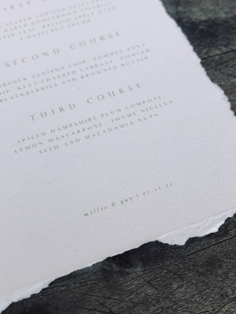 May include: A close-up of a white paper menu with a torn edge. The menu lists three courses with detailed descriptions of each dish. The text "millie & guy | 07.11.21" is printed at the bottom. The paper rests on a dark wooden surface.