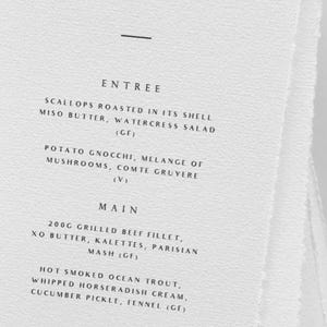 May include: A white menu card with the text "J & S MENU" at the top. The menu lists entree, main, and dessert options, including grilled beef fillet and wedding cake. The card has a simple, elegant design.