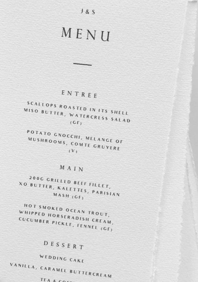 May include: A white menu card with the text "J & S MENU" at the top. The menu lists entree, main, and dessert options, including grilled beef fillet and wedding cake. The card has a simple, elegant design.
