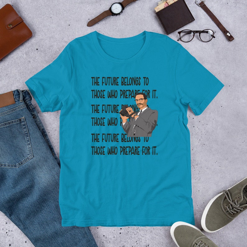 Puede incluir: Una camiseta azul turquesa con un estampado gr&aacute;fico negro de un hombre sosteniendo una c&aacute;mara. El texto en la camiseta dice "The future belongs to those who prepare for it."