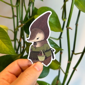 Op de afbeelding: Een grillige sticker met een cartoon egel karakter met een groene trui en een bruine hoed. De egel draagt een klein mandje met rode bessen. De sticker heeft een witte rand.