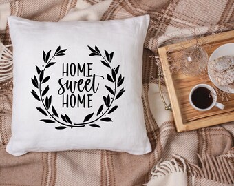 May include: White throw pillow with black text that reads "Home sweet home" surrounded by a wreath of leaves. The pillow is on a brown and white plaid blanket.