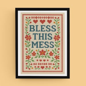 May include: Framed cross-stitch art with the words "BLESS THIS MESS" in blue, surrounded by red hearts, flowers, and green vines. The artwork is framed in a black frame.